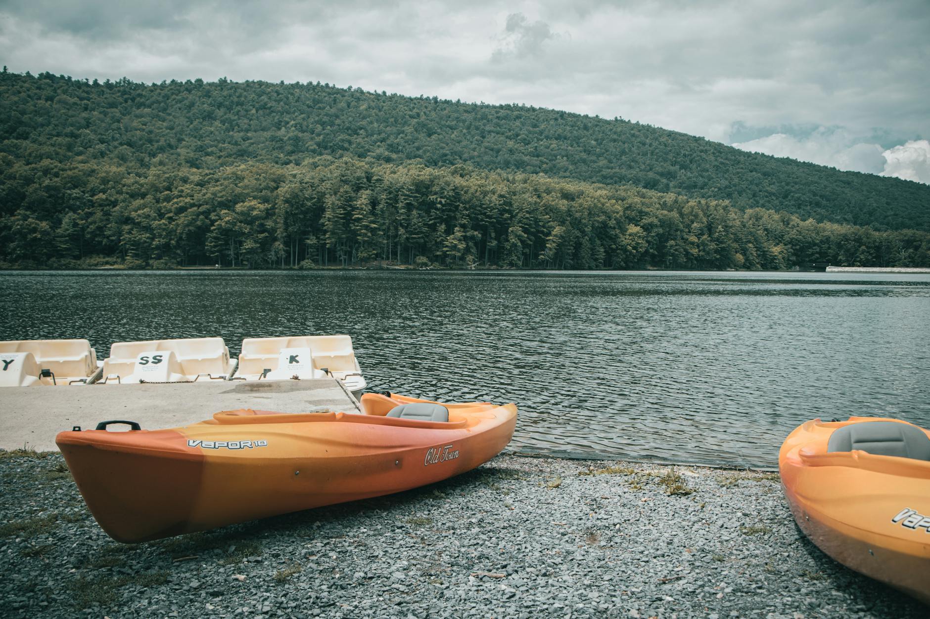 Peaceful lake scene with orange kayaks on the shore and a forested hillside.
