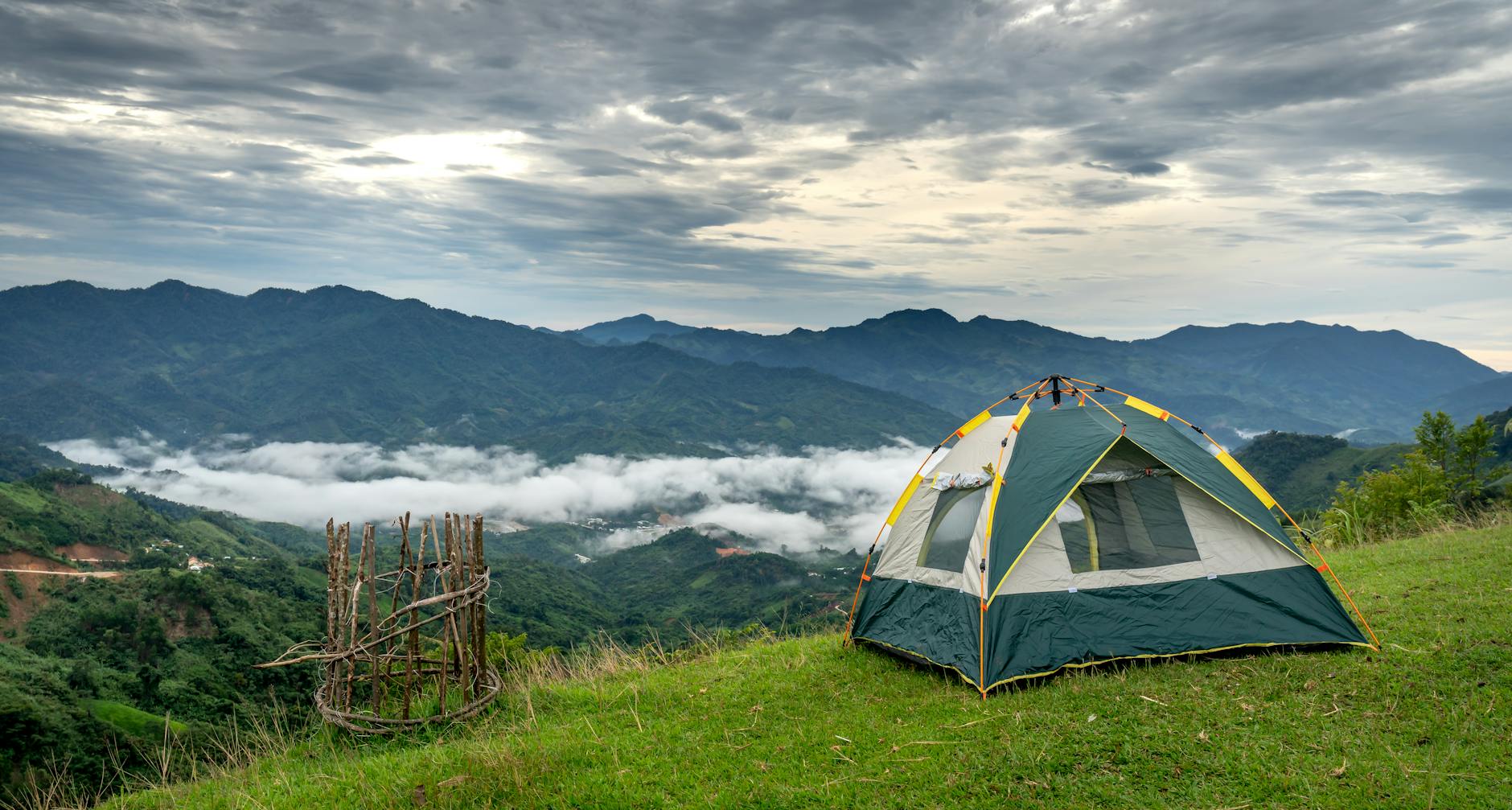 Picturesque mountain campsite with a tent, overlooking a dramatic cloudy sky and lush landscapes.