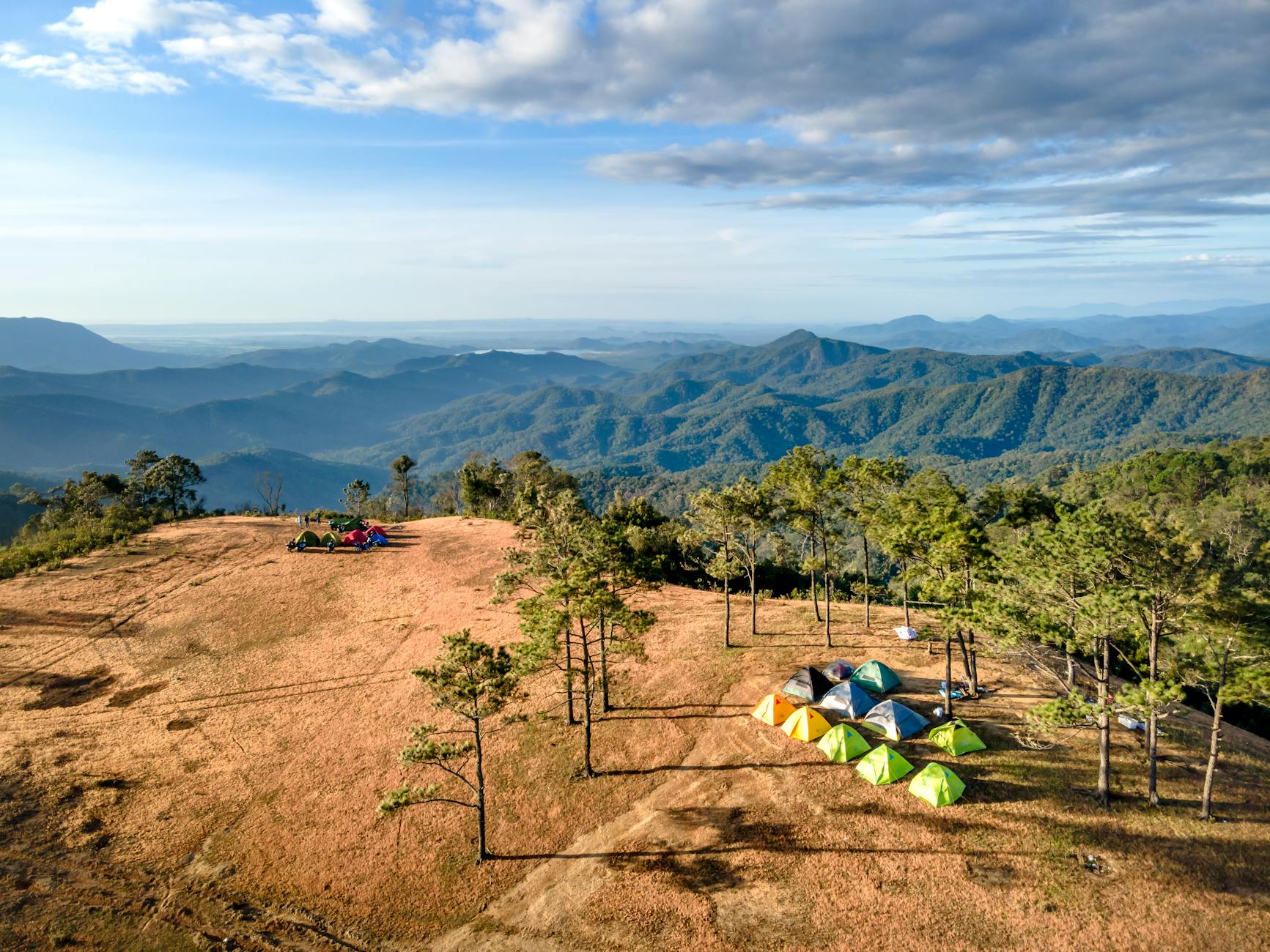Aerial shot of vibrant tents scattered on a hilltop overlooking a vast mountain range under a partly cloudy sky.