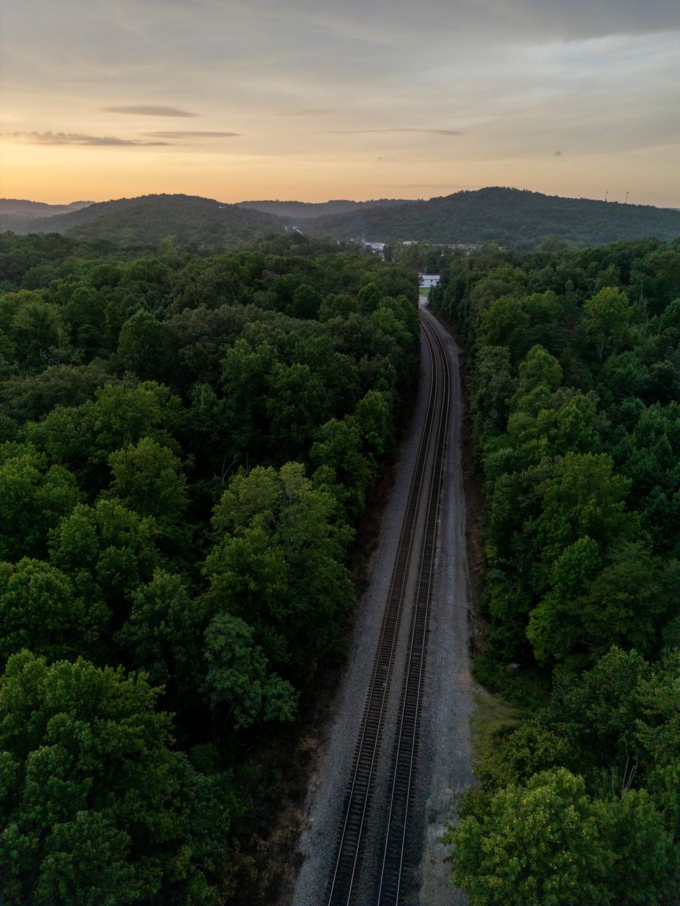 Stunning aerial view of a railroad cutting through dense forest with a scenic sunset backdrop.