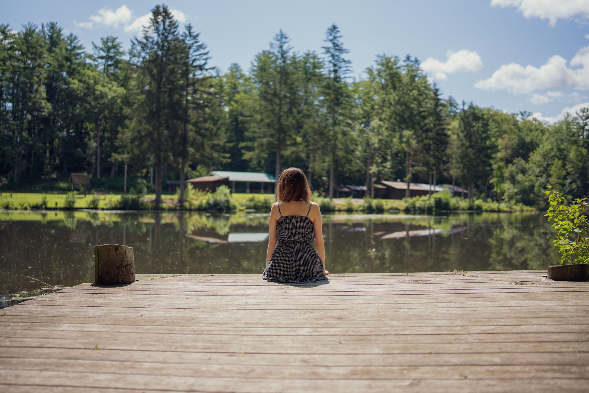 Serene view of a woman sitting on a wooden dock by a peaceful lakeside with lush trees in the countryside.